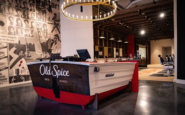 : An image showing the new Old Spice barbershop. The front desk is shaped like a boat, Old Spice ad images on the wall, and barbershop chairs fill the retail space.