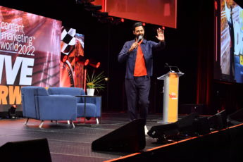 Brown skinned man with a goatee holding a microphone on stage. He's wearing an orange shirt with a blue shirt and dark pants.