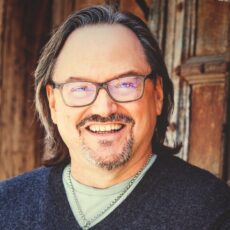 Robert Rose is wearing glasses, a grey shirt with a blue v-neck sweater over it, and has brown hair.