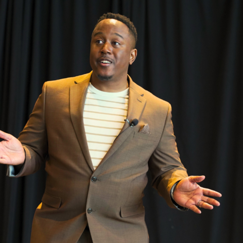 Troy Sandidge, founder of Strategy Hackers, gestures while wearing a brown jacket in front of a black background. 
